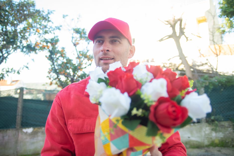 A male delivery person wearing a red jacket and matching red cap is holding a vibrant bouquet of fresh flowers, primarily consisting of red and white roses, wrapped in colorful, patterned paper. The b