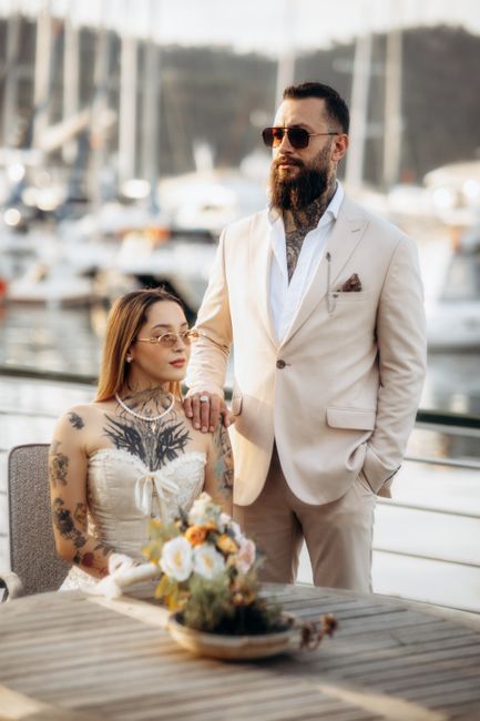A man and woman are photographed outdoors near a marina with boats docked in the background, likely during a wedding or celebration near Erith Pier and Thames. The woman is seated at a table, wearing 