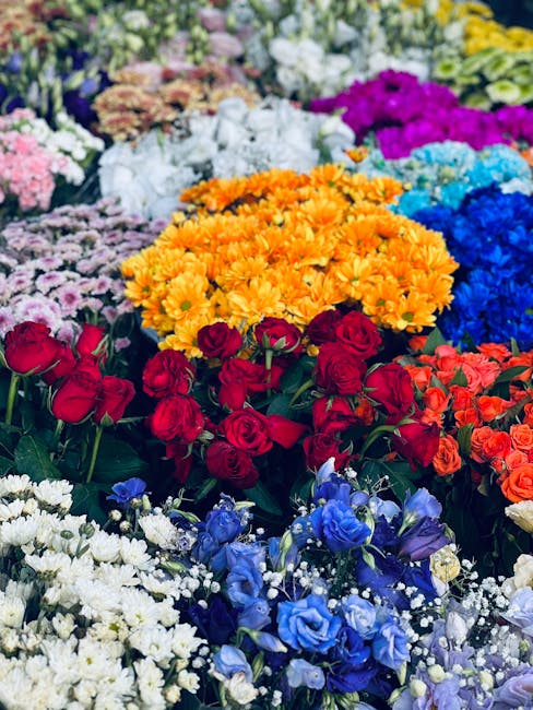 A vibrant floral display featuring an assortment of fresh flowers arranged in a dense, colorful bouquet. Prominent in the foreground are clusters of deep red roses with velvety petals, surrounded by b
